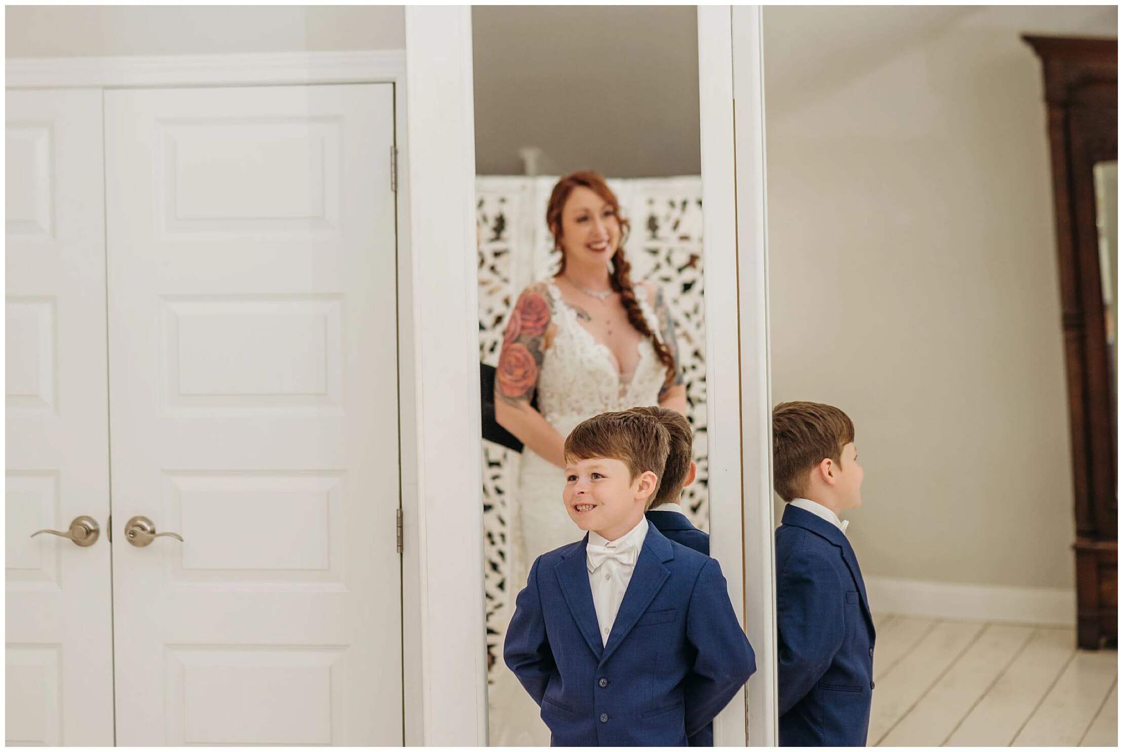 young son leaning against a mirror and watching his mom in her wedding dress