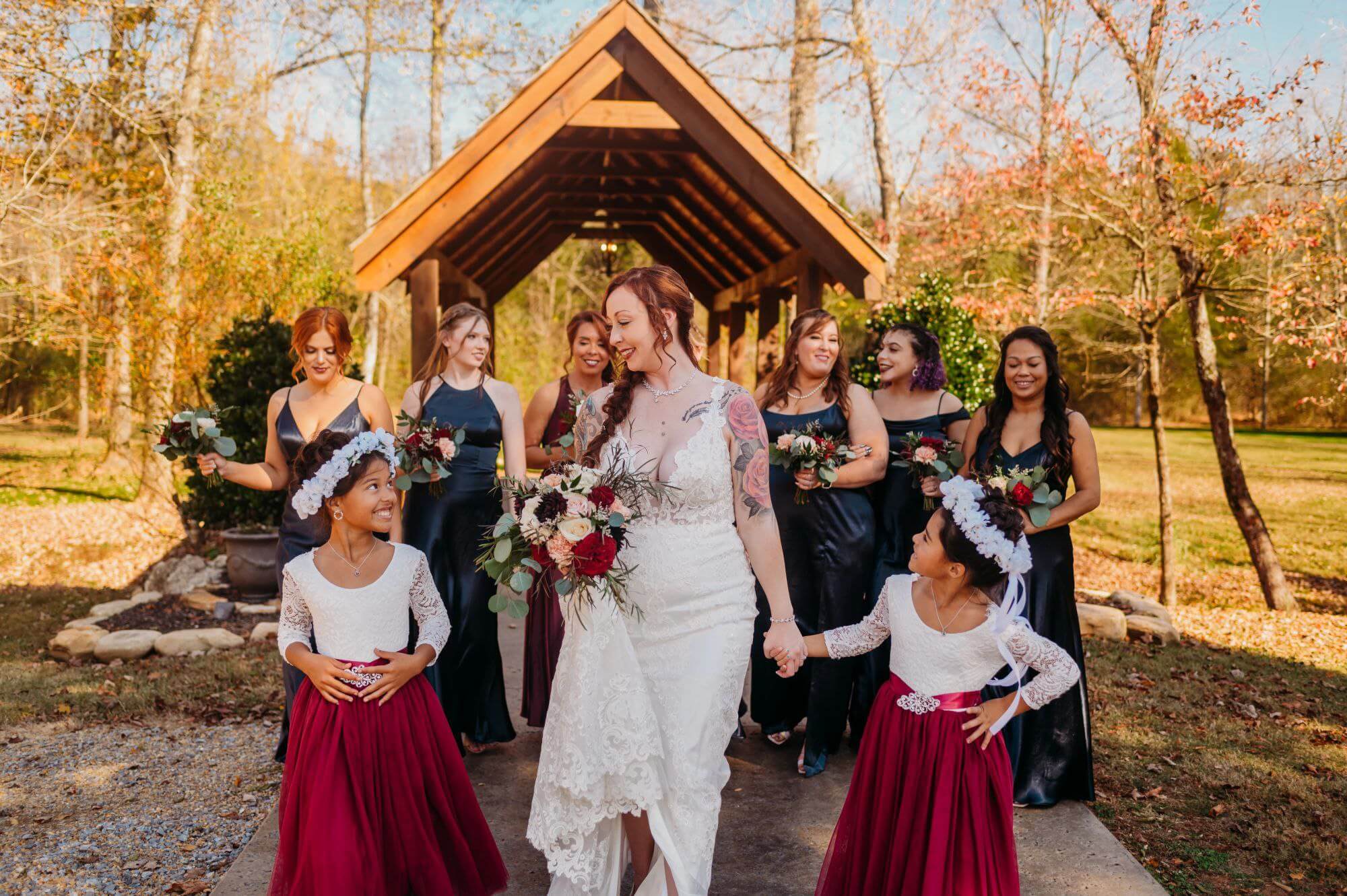 bride walking with flower girls in front of bridesmaids and a wooden bridge