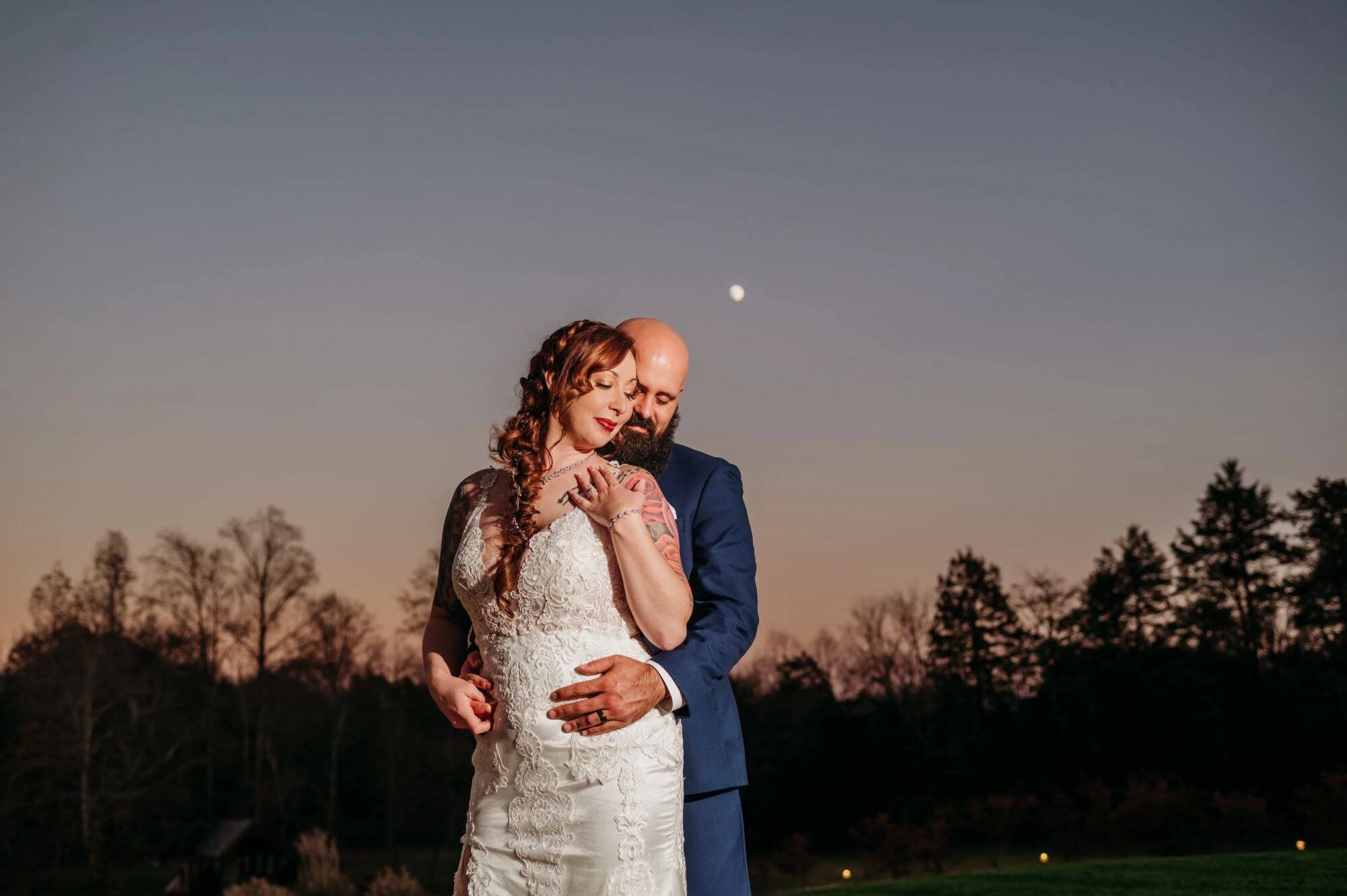 groom hugging bride's back at dusk with the moon showing in the background