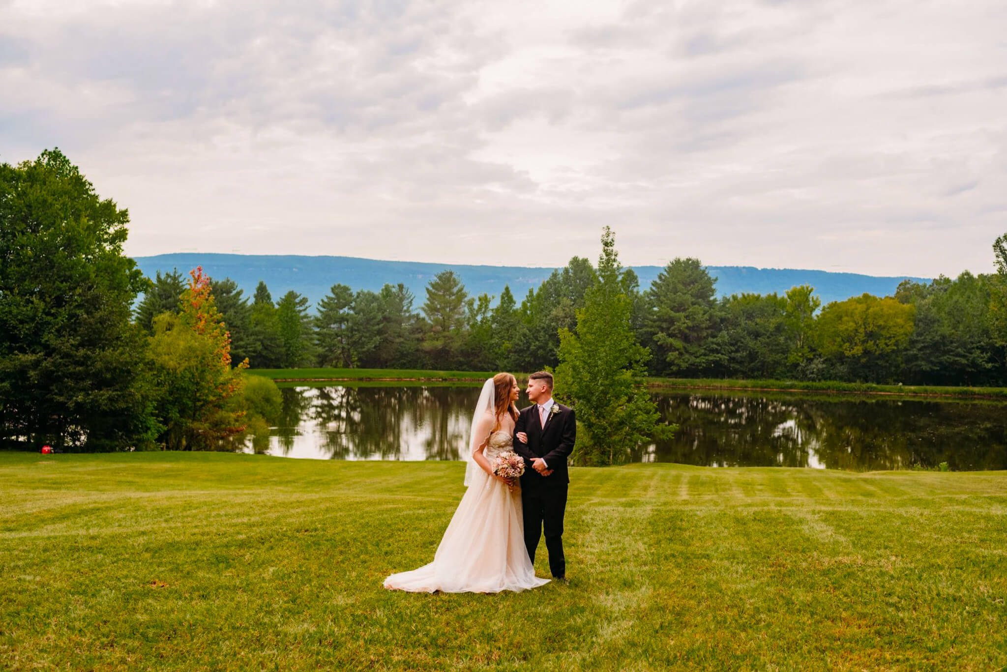 photo of a bride hugging a groom's arm with a pond and mountains in the background