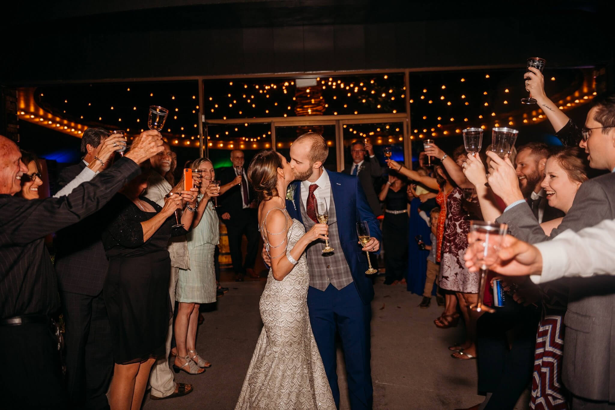 photo of bride and groom kissing with guests raising their glasses in a toast and bistro lights in the background